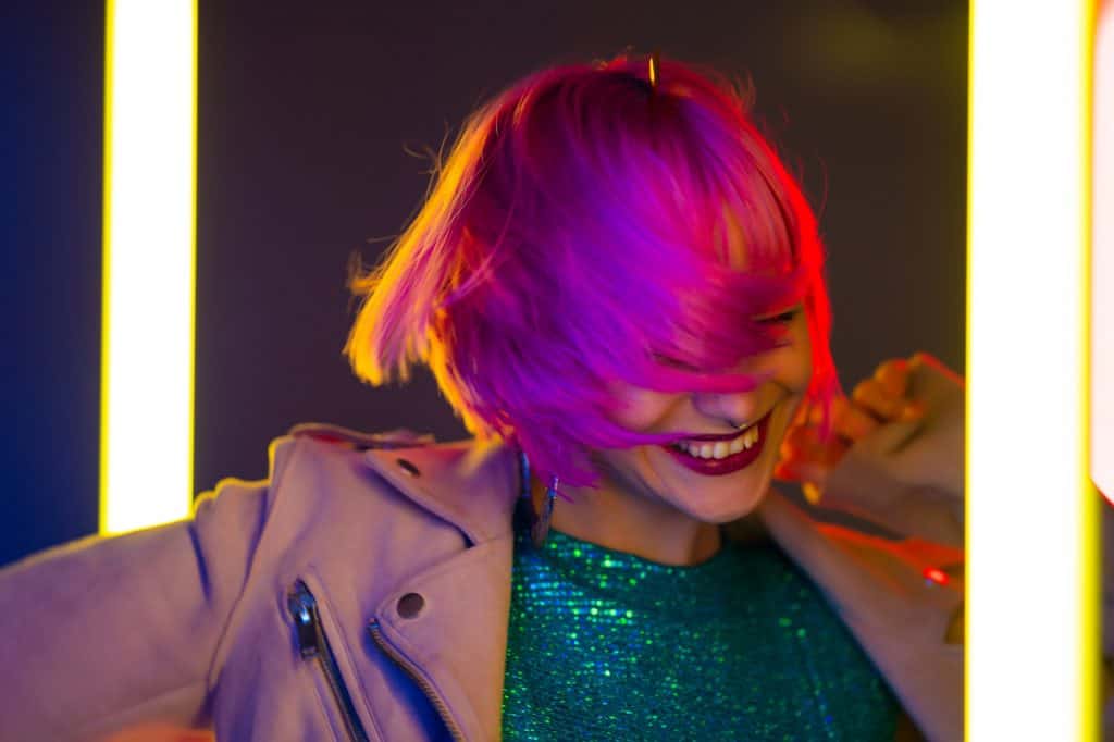 Amazing smiling woman with dyed pink hairstyle dancing on led-colorful neon lamps background.